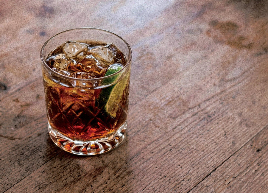 Rambler Libre cocktail made with Rambler Campfire Spiced Rum, served over ice with a lime wedge in a crystal tumbler on a wooden table